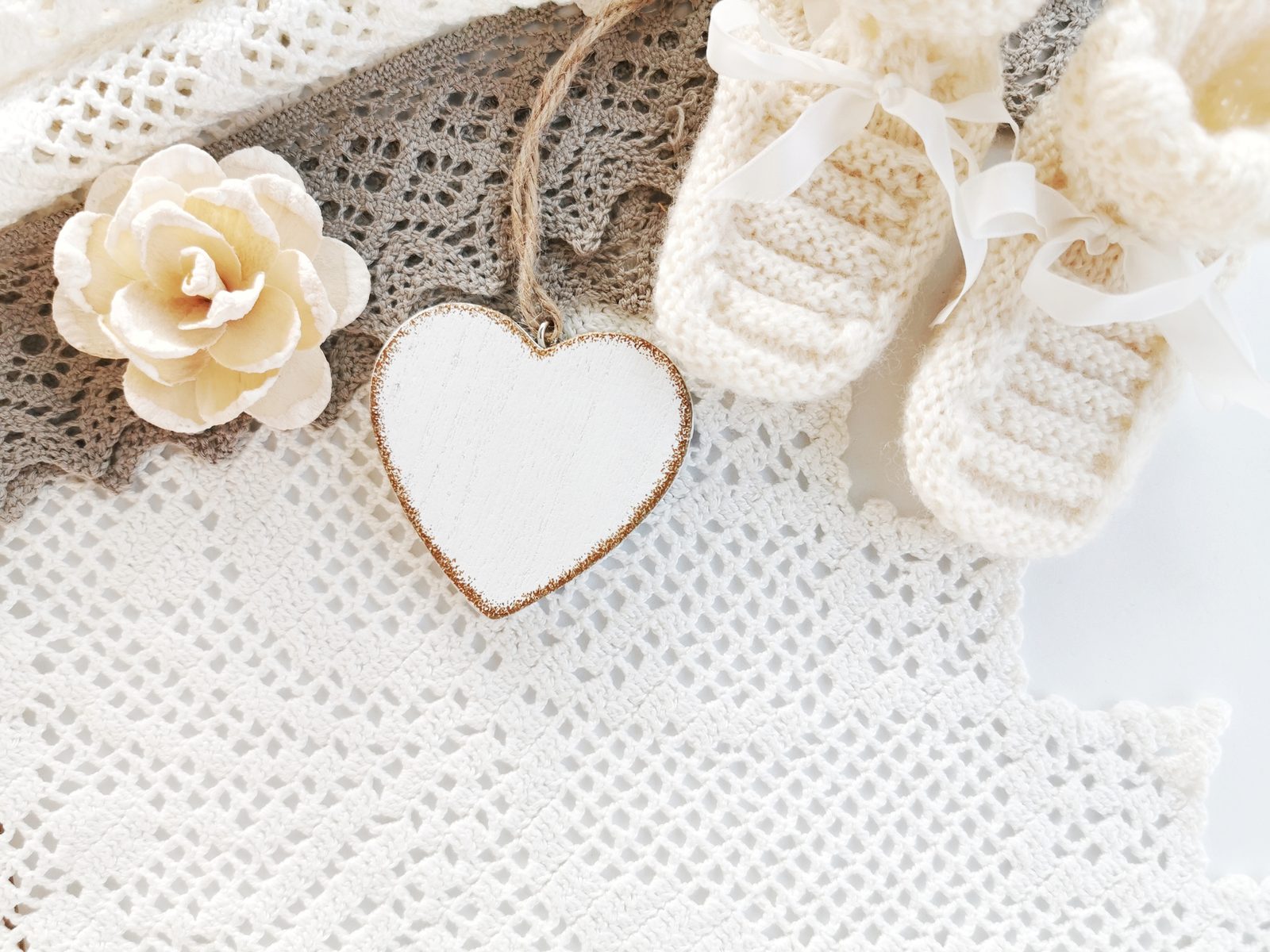 White knitted baby booties, a white wooden heart and a cream paper rose on lace.