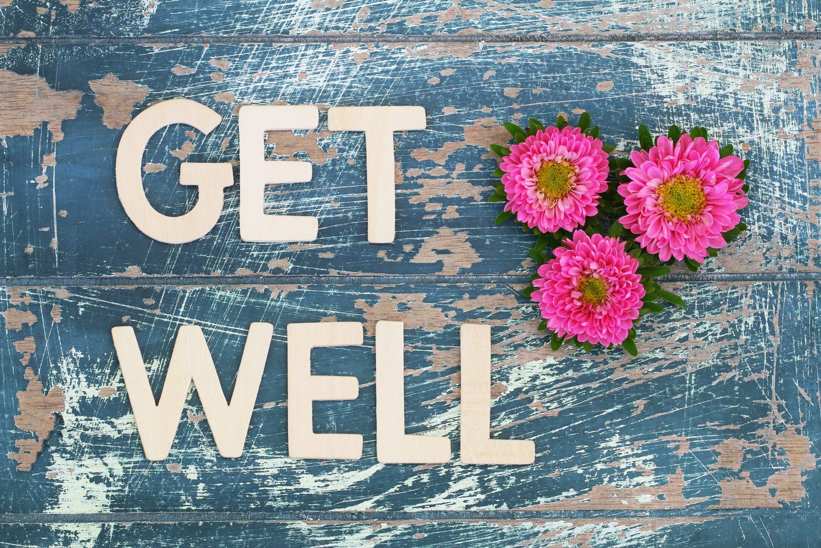 Wooden letters spelling GET WELL on weathered blue boards, with three pink aster flowers.