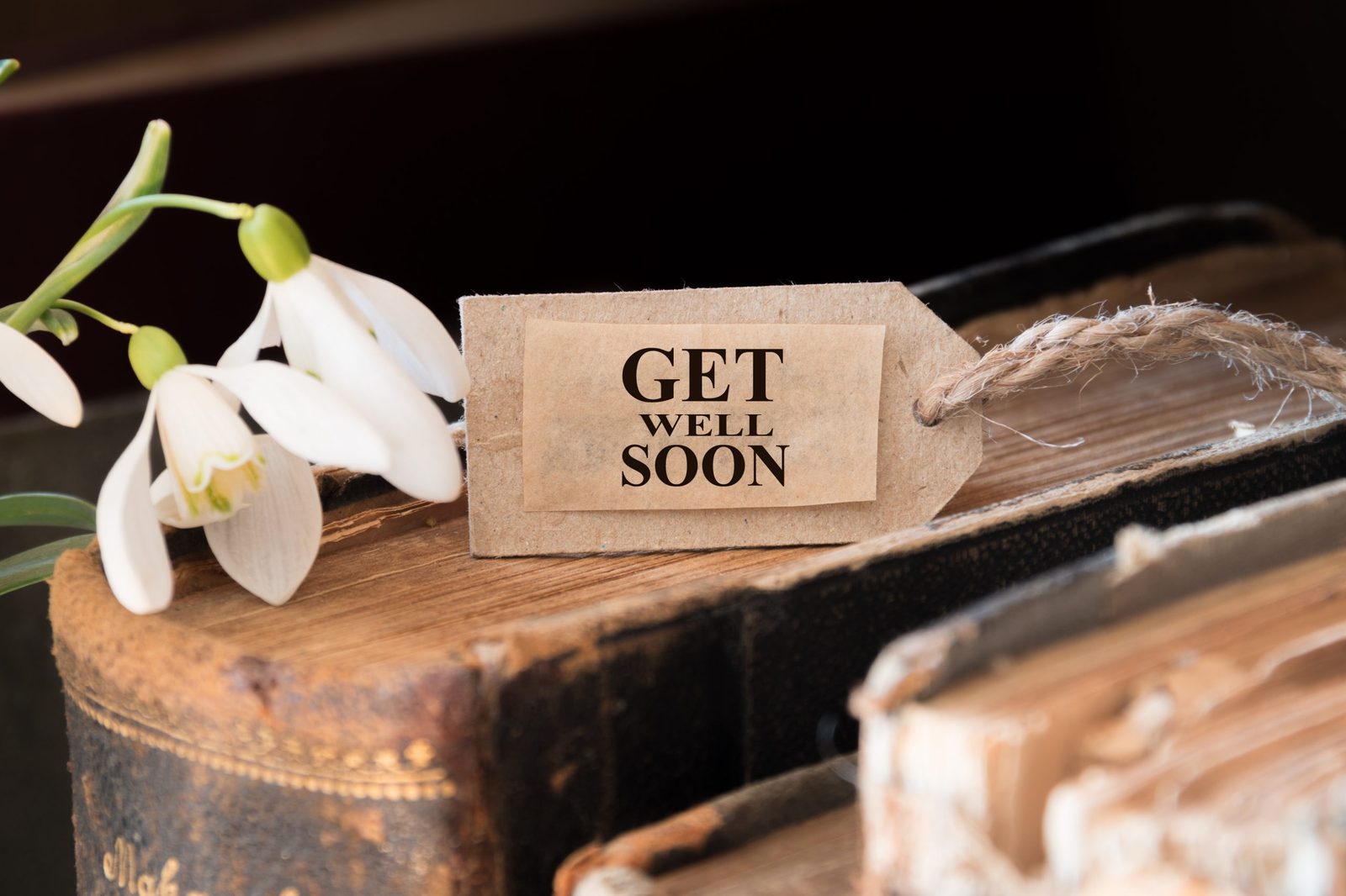 A paper tag reading GET WELL SOON beside snowdrop flowers on old books.