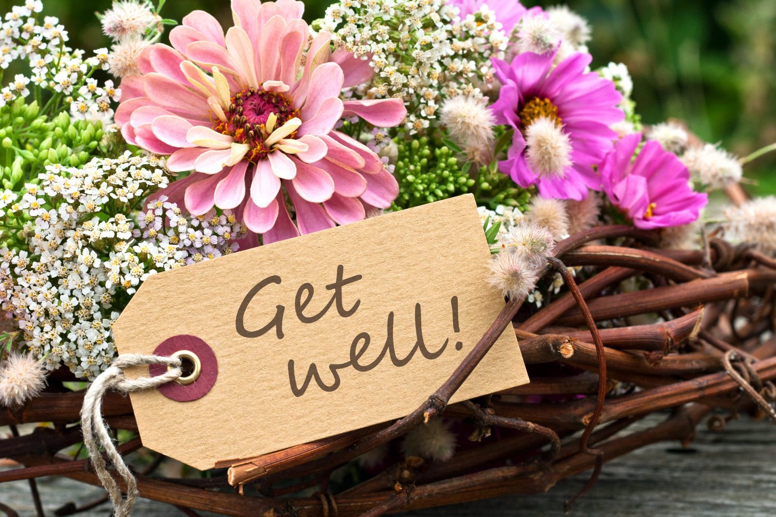 A kraft paper tag reading Get well! on a wreath of twigs, with pink and purple flowers.