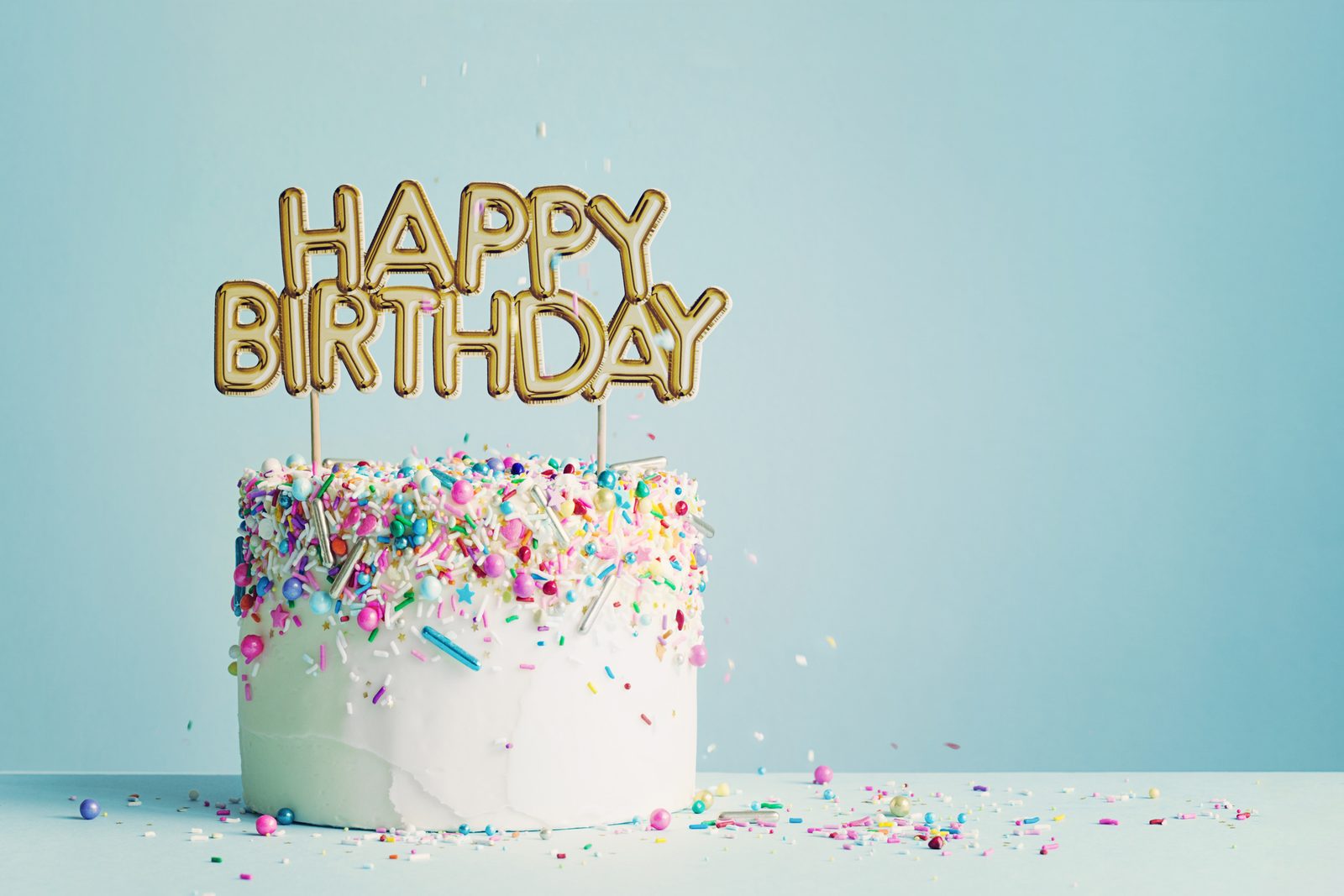 A frosted cake with rainbow sprinkles, topped with gold HAPPY BIRTHDAY letters, against a pale blue background.